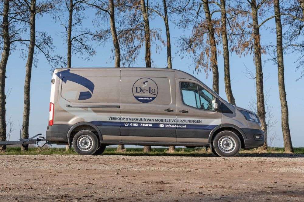 De Lo bedrijfsbus geparkeerd op een landelijke locatie met bomen en een heldere blauwe lucht op de achtergrond.