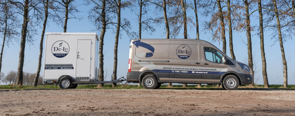 De Lo bedrijfsbus met aanhanger waarop een mobiele badkamer is bevestigd, geparkeerd op een landelijke locatie met bomen en een heldere blauwe lucht op de achtergrond.