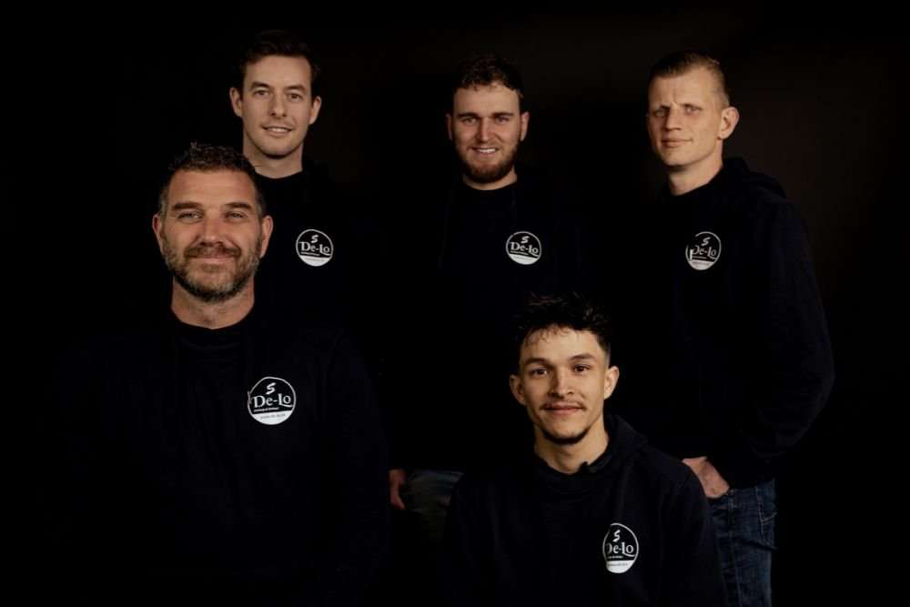 Groepsfoto van vijf medewerkers van De Lo, gekleed in donkere hoodies met het bedrijfslogo, poserend tegen een zwarte achtergrond.