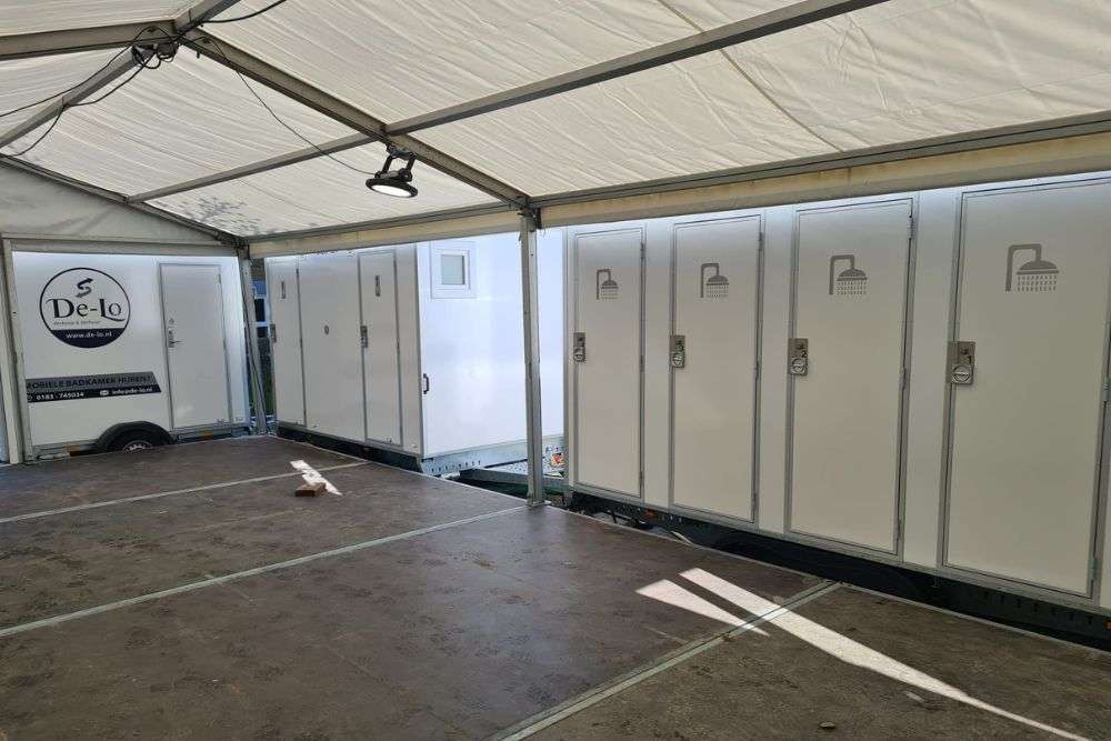 Mobiele douche units van De Lo opgesteld in een overdekte ruimte, met een aanhanger waarop 'Mobiele badkamer huren' staat vermeld.
