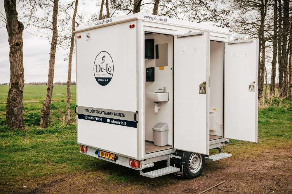 Vacuum toiletwagen van De Lo met geopende deuren, voorzien van een wastafel, spiegel en afgesloten toiletruimtes, geplaatst in een landelijke omgeving.